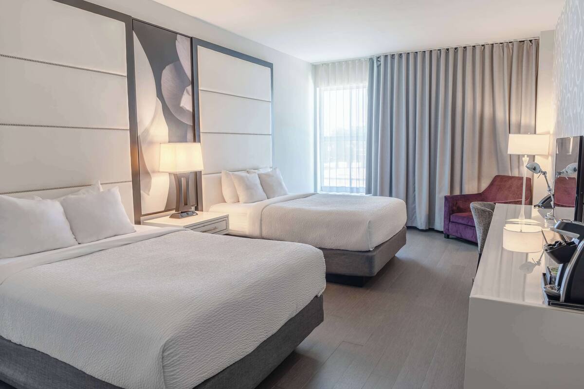 A hotel room with two beds, a desk, a chair, and a large window with curtains.