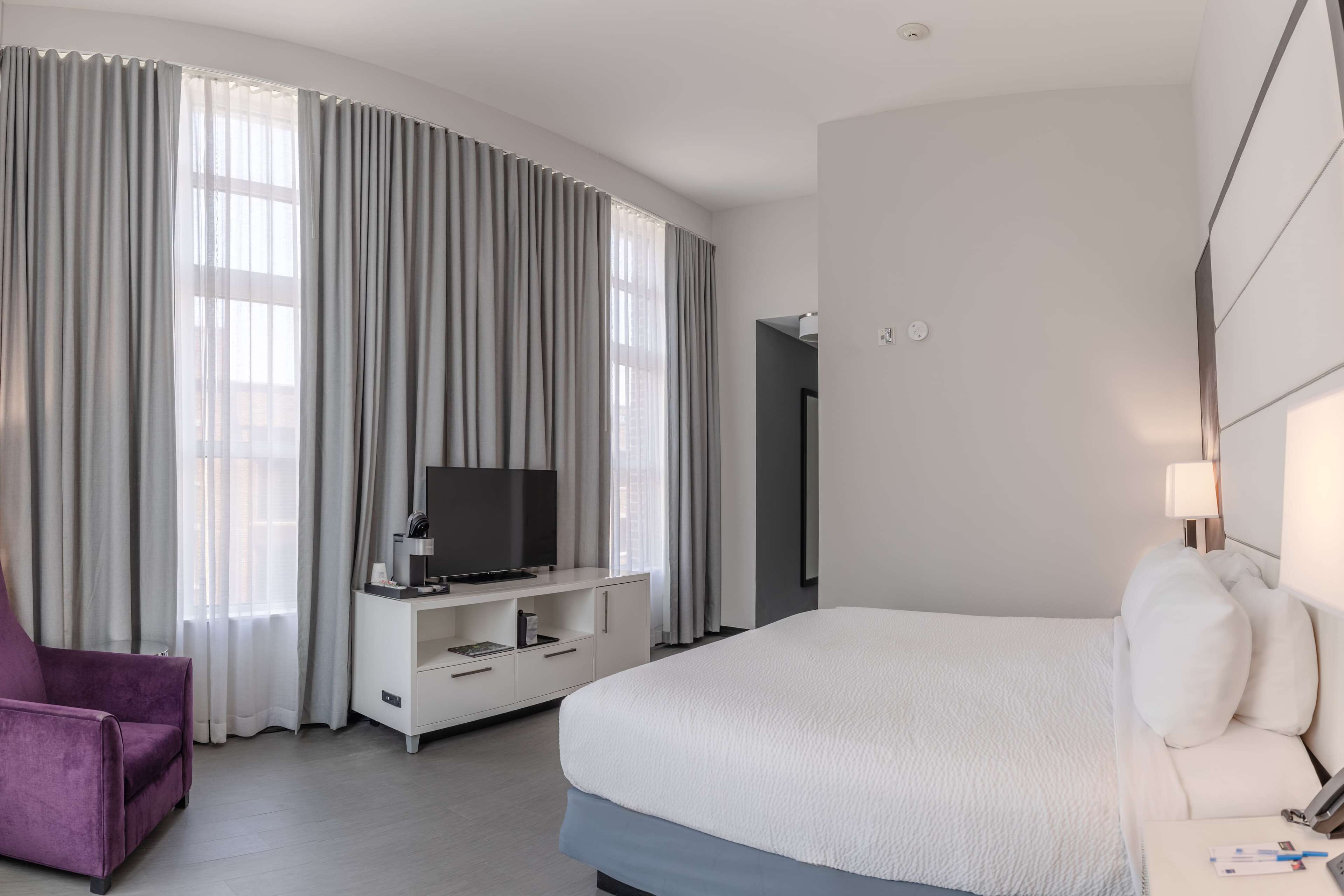A modern hotel room with a large bed, a TV on a white console, a purple armchair, and a large window with curtains.