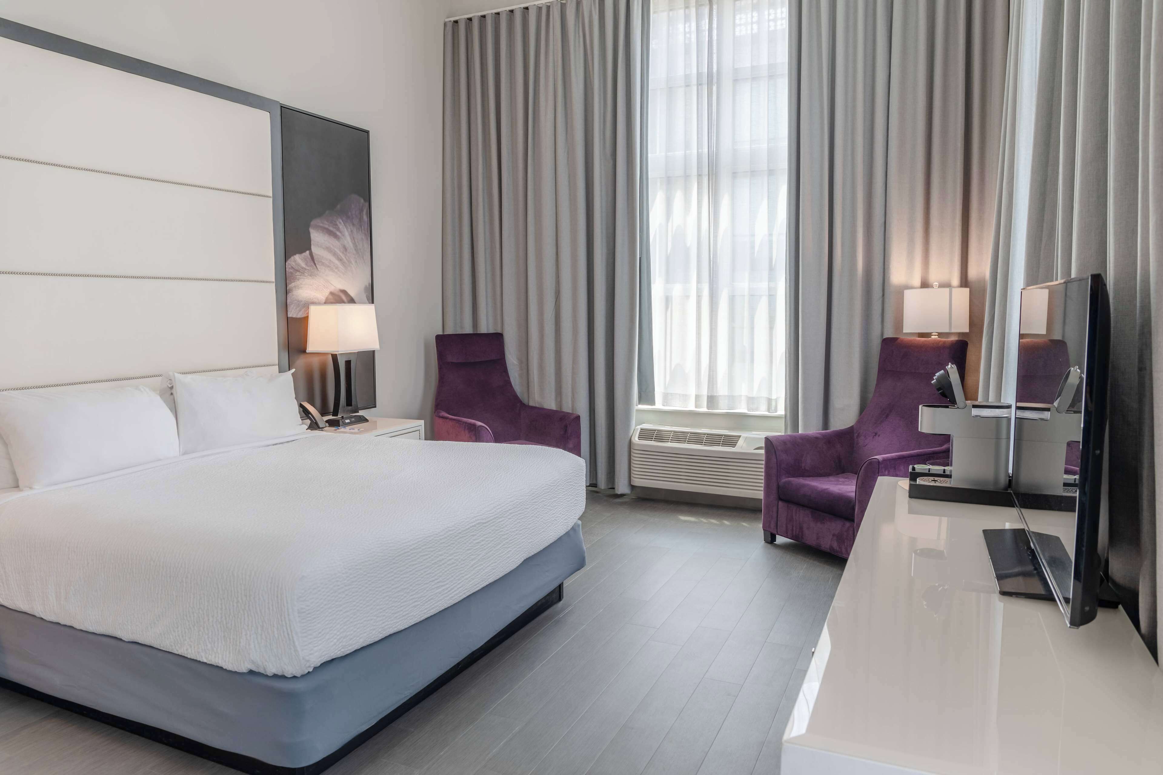 A hotel room with a large bed, two purple armchairs, a desk with a flat-screen TV, and a large window with curtains.