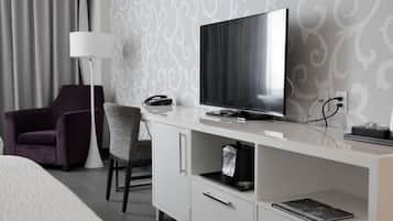 A modern hotel room with a flat-screen TV on a white console, a purple armchair, a floor lamp, and a patterned wallpaper.