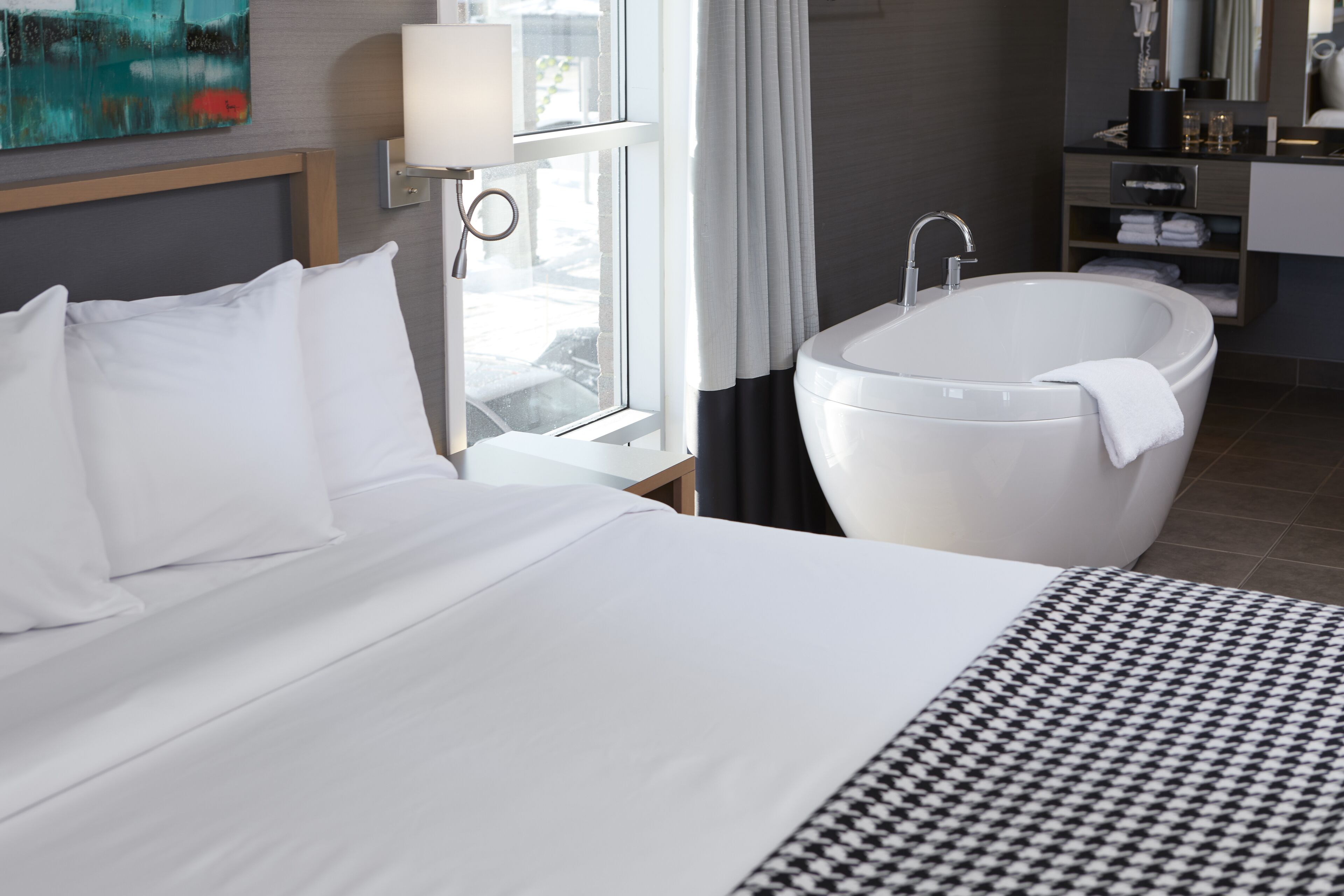 A hotel room with a large bed, a white bathtub, a window with curtains, and a bathroom with a mirror and shelves.