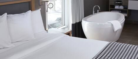A hotel room with a large bed, a white bathtub, a window with curtains, and a bathroom with a mirror and shelves.