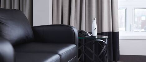 A modern hotel room with a black leather sofa, a glass side table with a telephone and a water bottle, and a window with curtains.