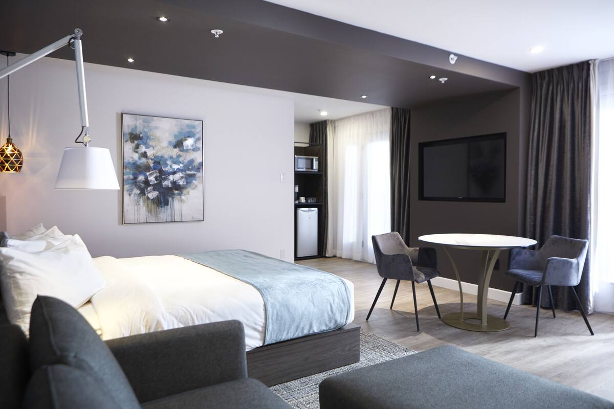 A modern hotel room with a large bed, a dining table with chairs, a flat-screen TV, and a painting on the wall.