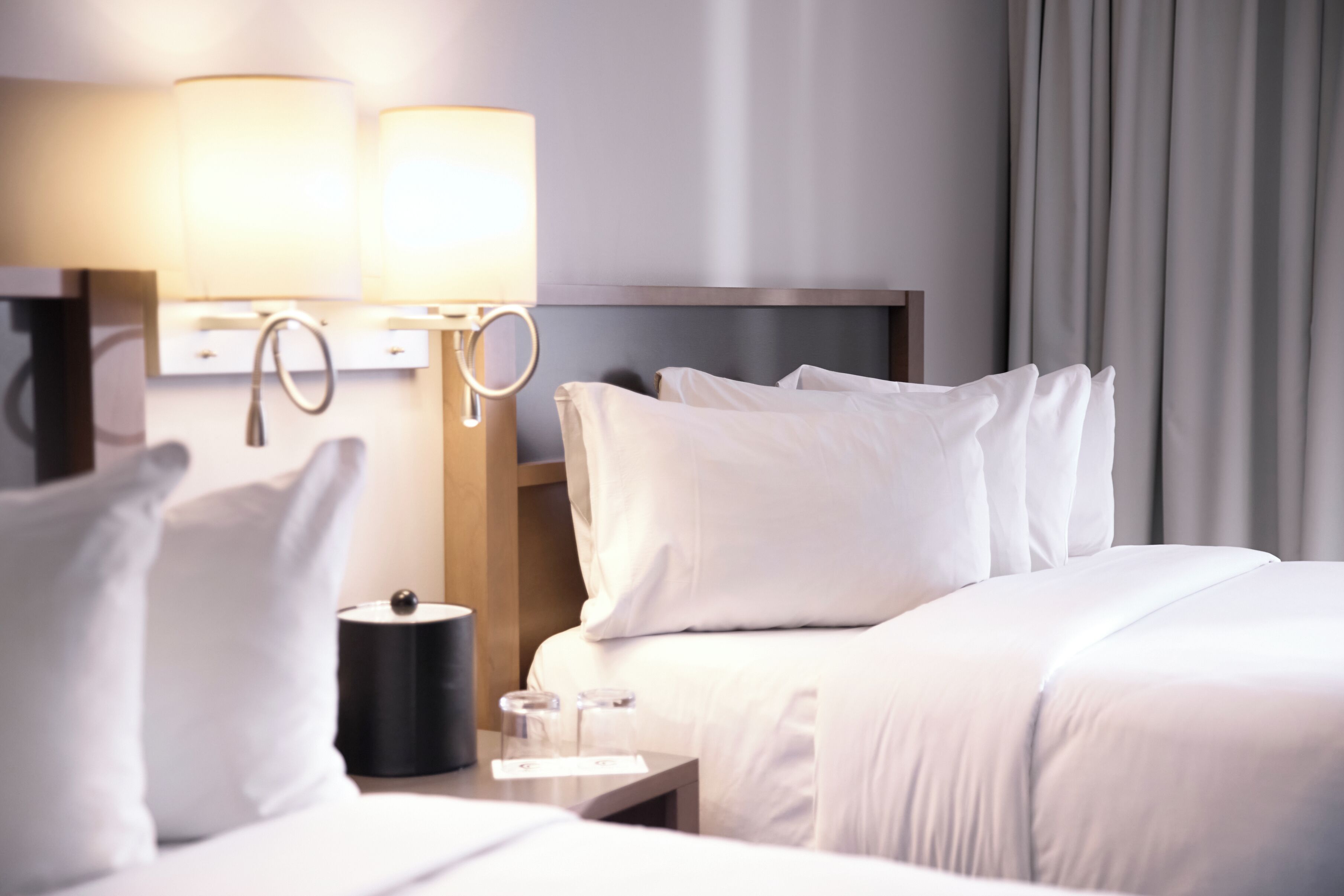 A hotel room with a neatly made bed, two bedside lamps, a small table with a lamp, and a window with curtains.