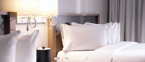 A hotel room with a neatly made bed, two bedside lamps, a small table with a lamp, and a window with curtains.