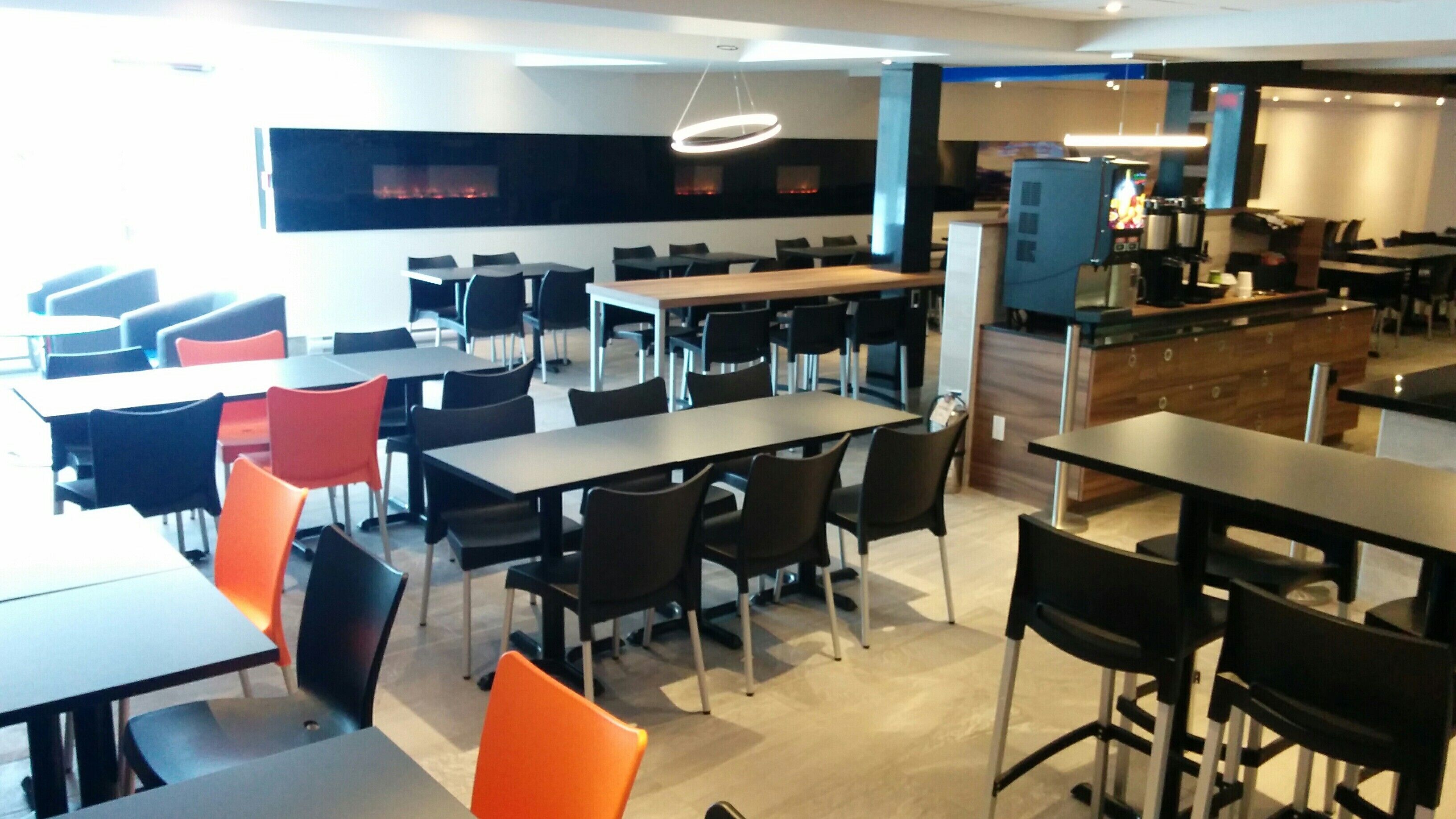 A modern dining area with multiple tables and chairs, a kitchen area with a counter and appliances, and a large window with a view of the outside.