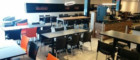 A modern dining area with multiple tables and chairs, a kitchen area with a counter and appliances, and a large window with a view of the outside.