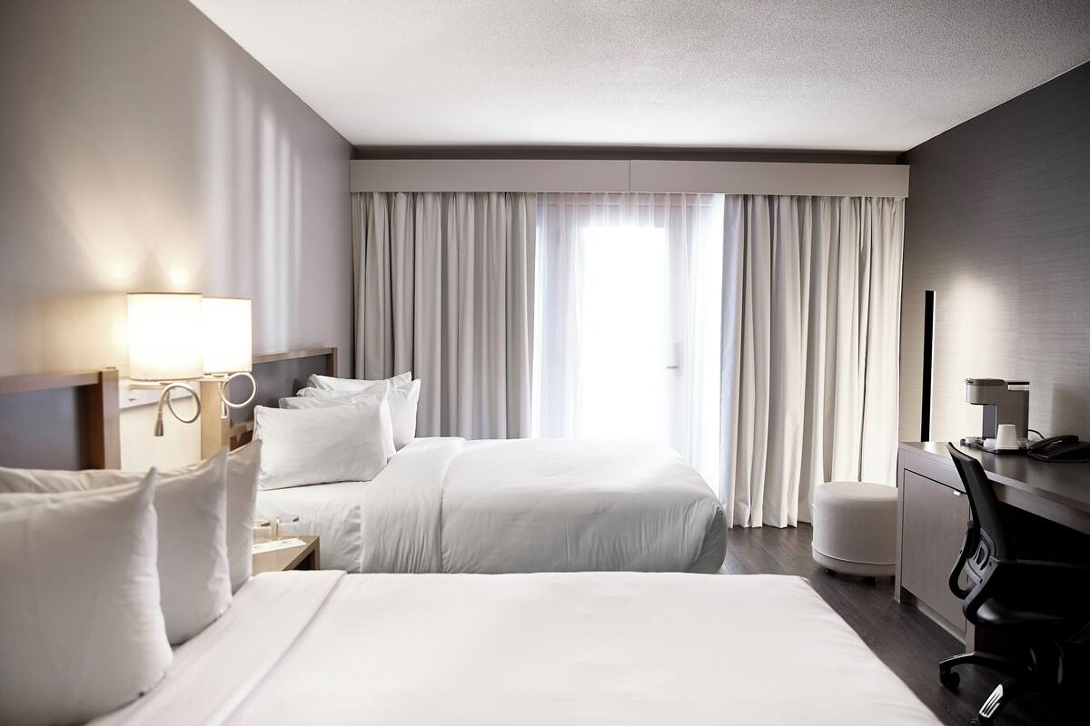A hotel room with two beds, a desk, a chair, a lamp, and a large window with curtains.