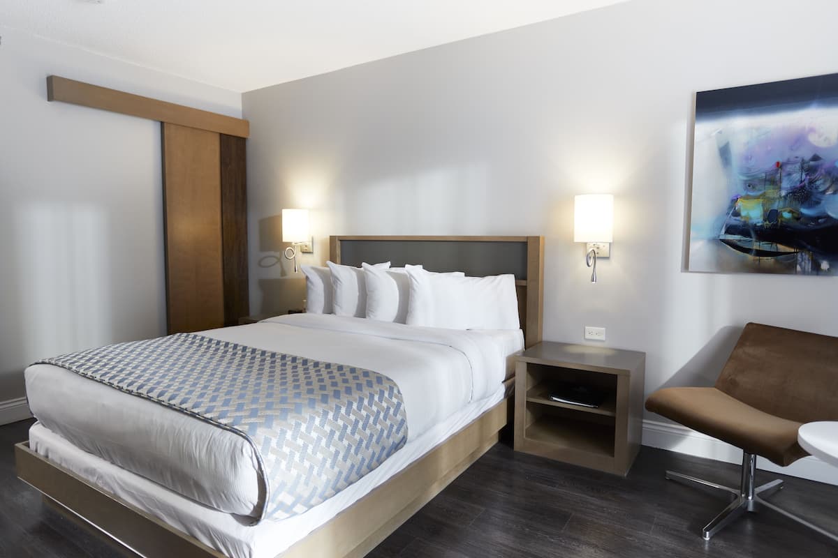 A modern hotel room with a large bed, bedside tables, a chair, and a painting on the wall.