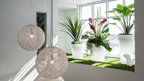 A modern interior with spherical hanging lights, potted plants, and a view of a cityscape through large windows. - Hôtel Cofortel (L'Ancienne-Lorette)