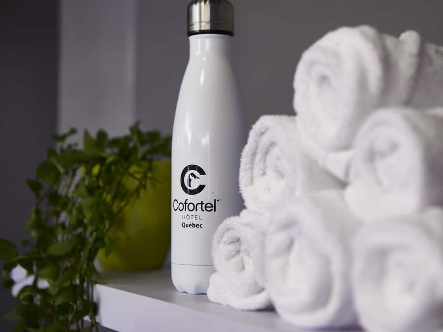 Una botella blanca con la etiqueta "Cofortel Hôtel Québec" y toallas blancas enrolladas cuidadosamente sobre una superficie blanca.