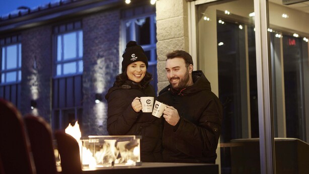 Two people holding mugs at an outdoor seating area with string lights and a fire feature. - Hôtel Cofortel (L'Ancienne-Lorette)
