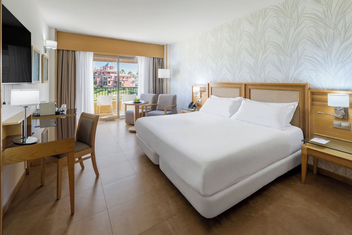 A hotel room with a large bed, a desk with a chair, a small table, and a view of buildings through the window.