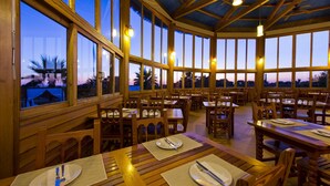 A spacious dining area with wooden tables and chairs, large windows offering views of palm trees and a sunset. - Elba Costa Ballena Beach & Thalasso Resort (Rota)