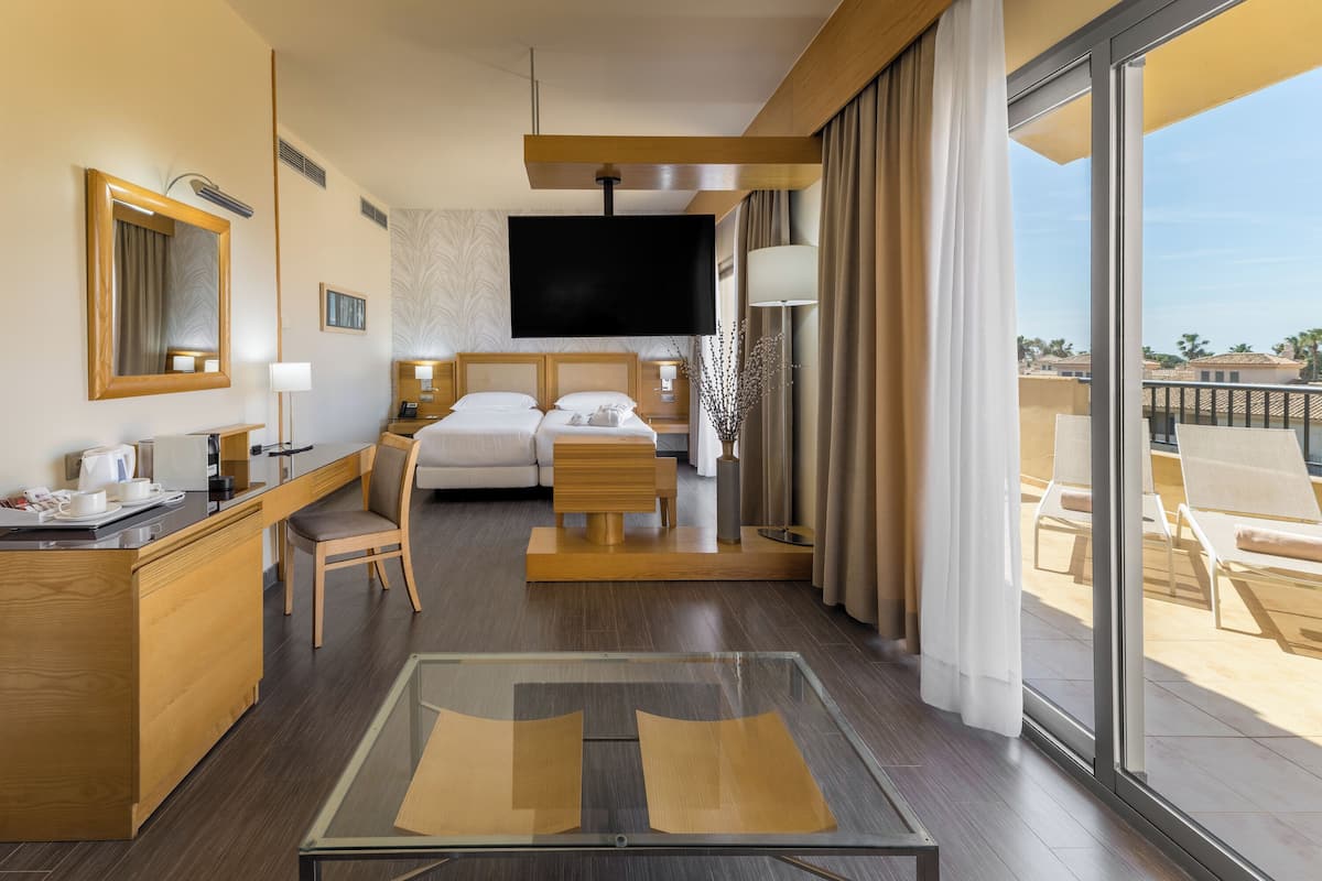 A hotel room with a bed, a desk, a TV, a balcony with chairs, and a large window.