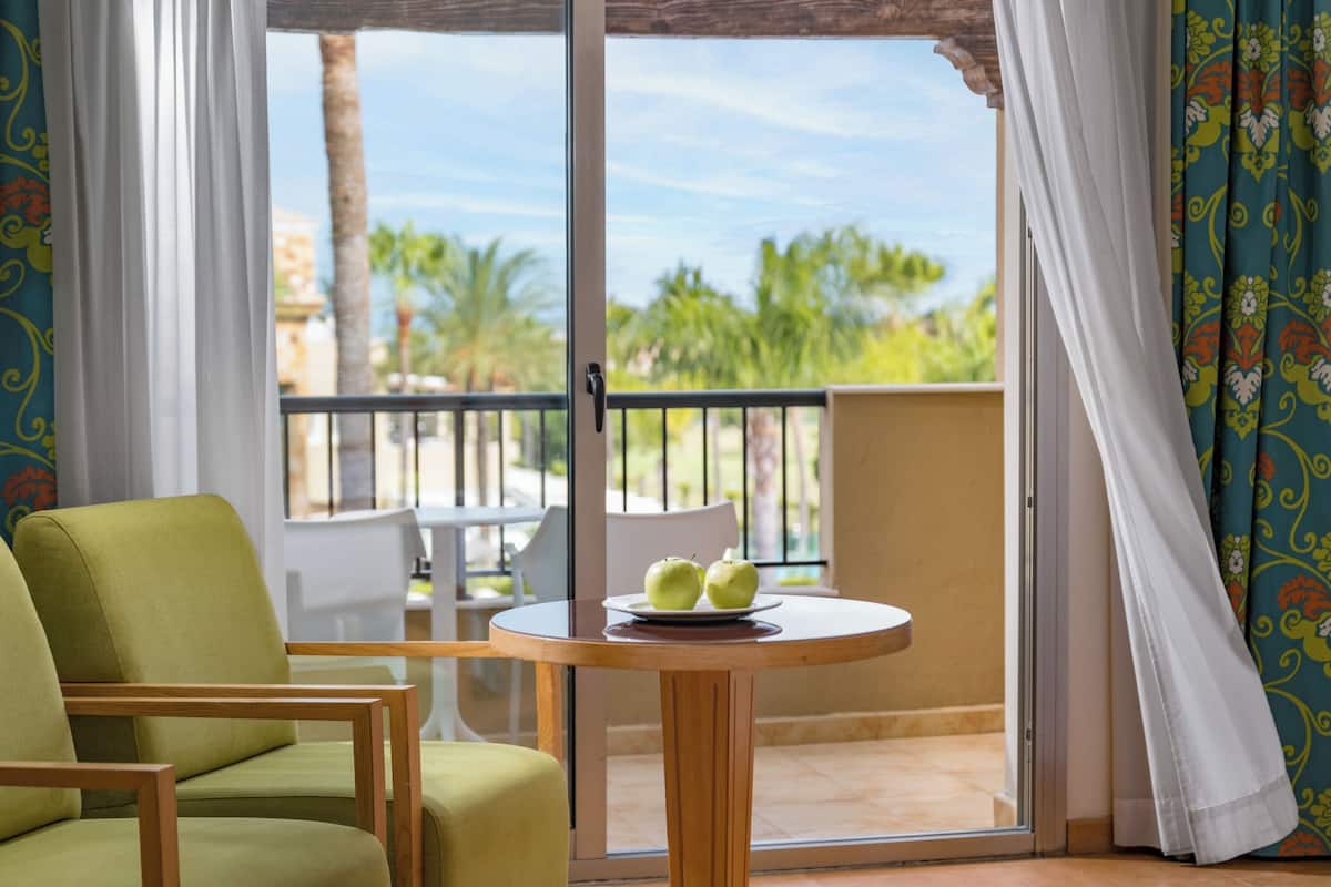 A room with a green chair, a table with two apples, and a view of palm trees and buildings through a sliding glass door.