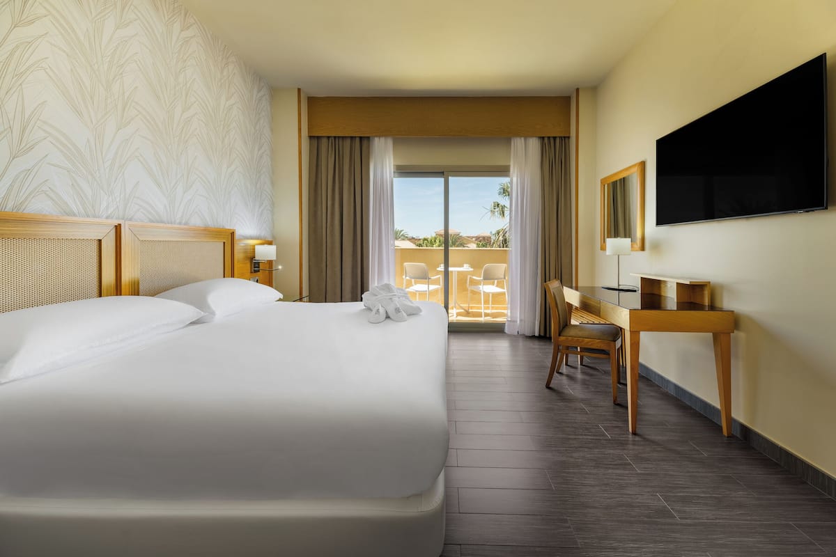 A hotel room with a large bed, a desk, a chair, a TV, and a balcony with a view.