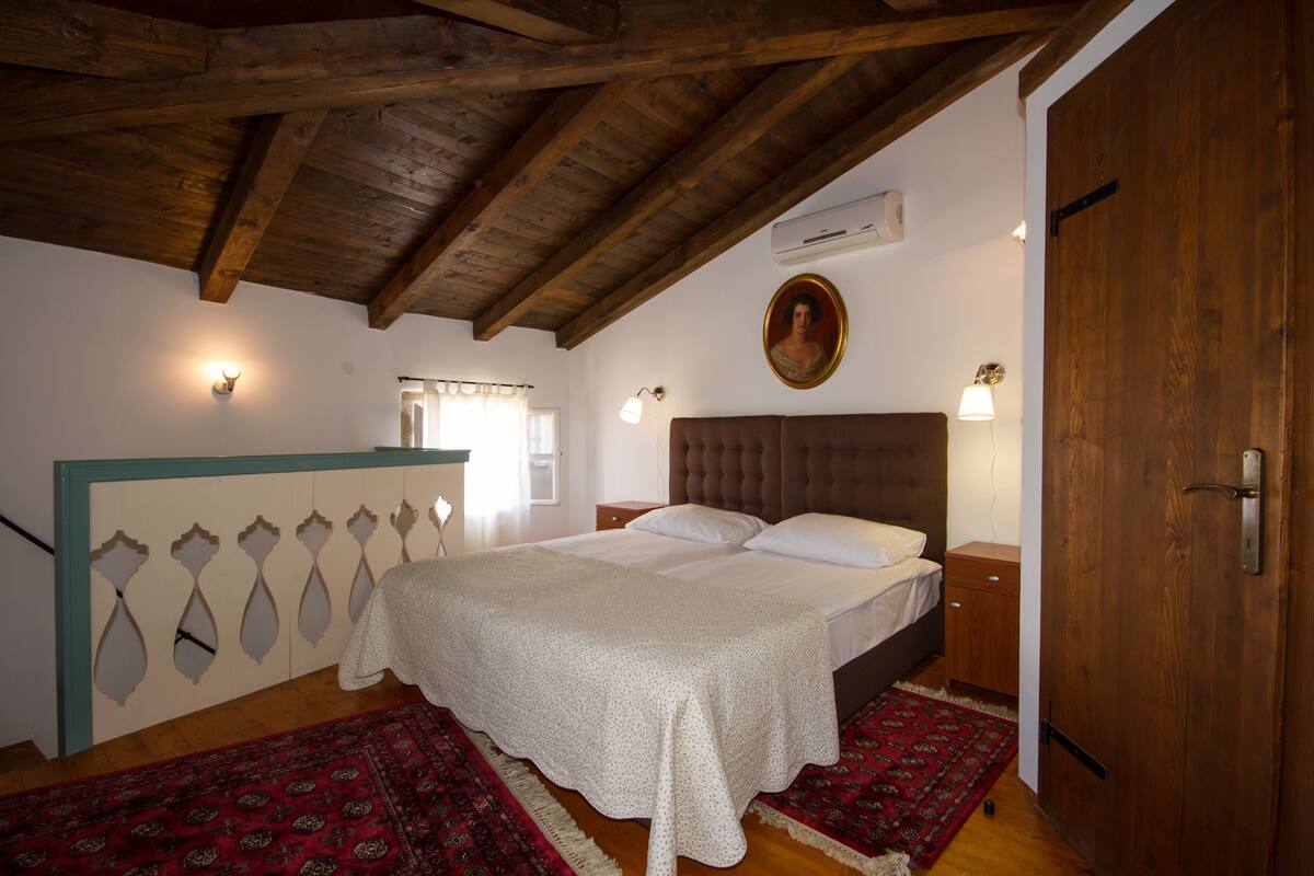 Een slaapkamer met een houten plafond, een bed met witte lakens, een rood geweven vloerkleed en een portret aan de muur.