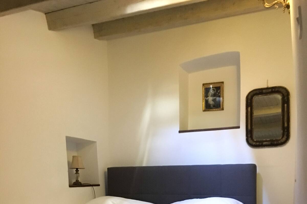 Een bed met een donker hoofdbord, een bedlampje, een ingelijst schilderij aan de muur en een spiegel.