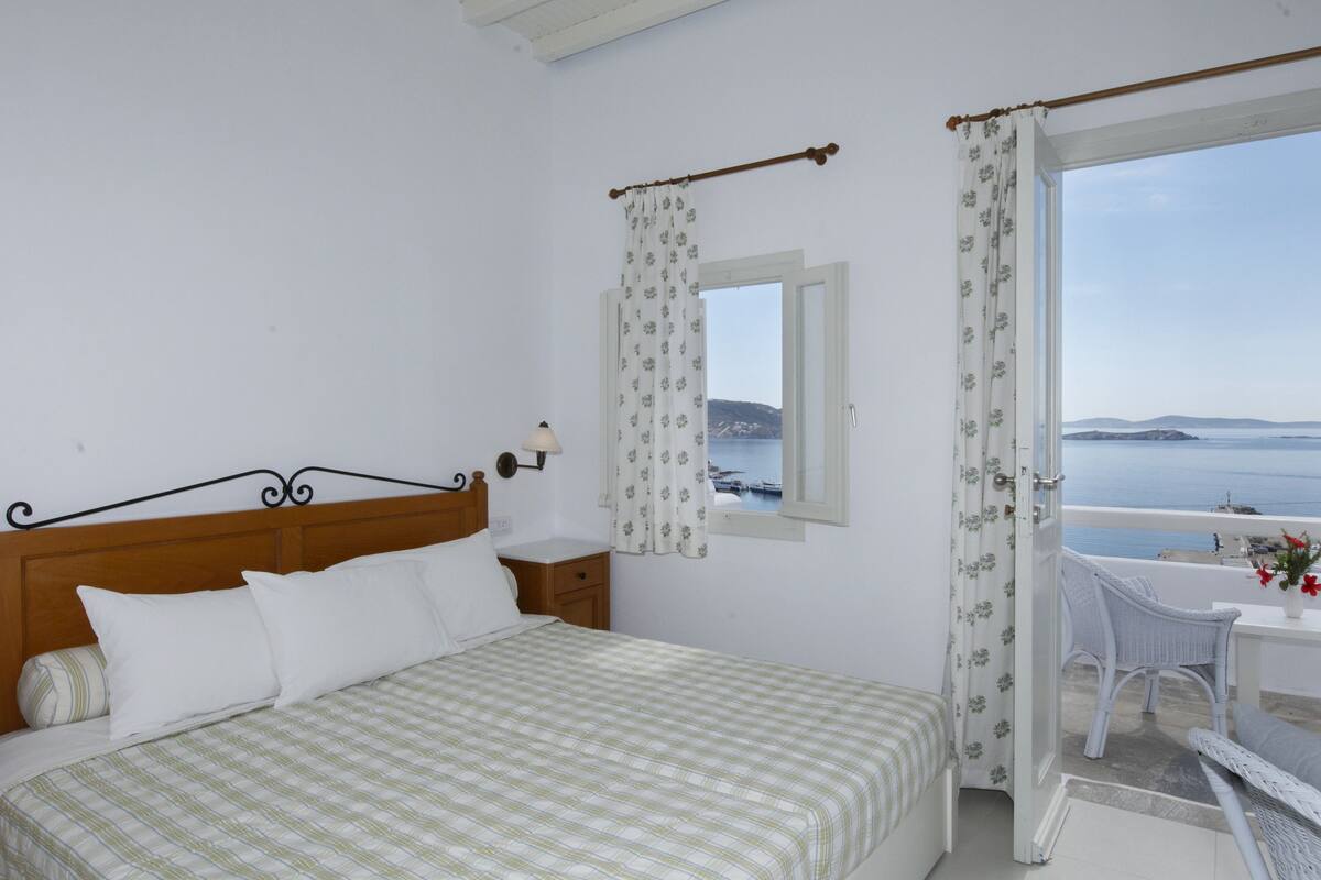 Un dormitorio con cama, mesita de noche, ventana con cortinas, balcón con una silla y vista al mar.