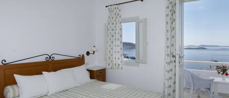 Un dormitorio con cama, mesita de noche, ventana con cortinas, balcón con una silla y vista al mar.