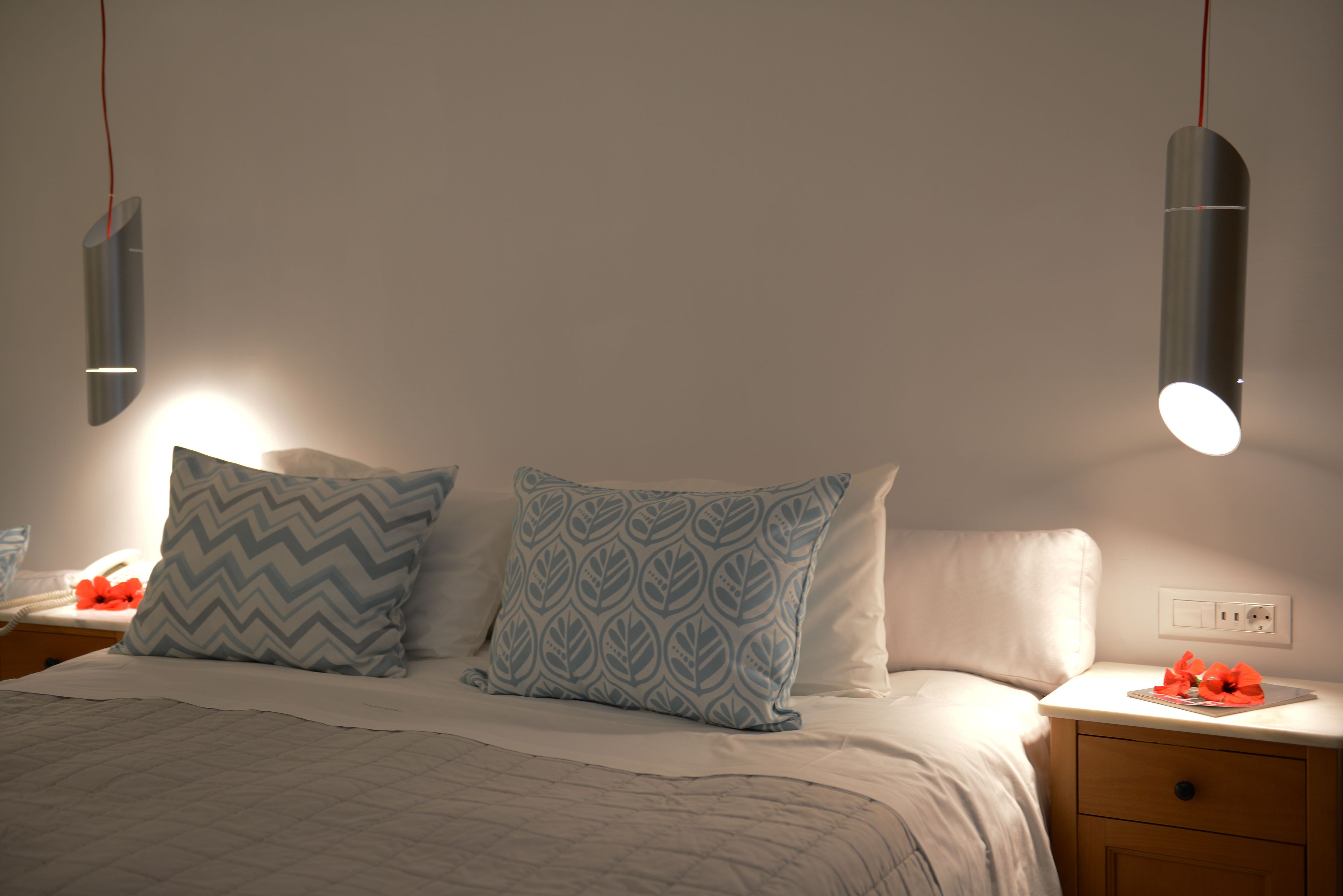 Een net opgemaakt bed met gestreepte kussens, een nachtkastje met een lamp en een stopcontact aan de muur.