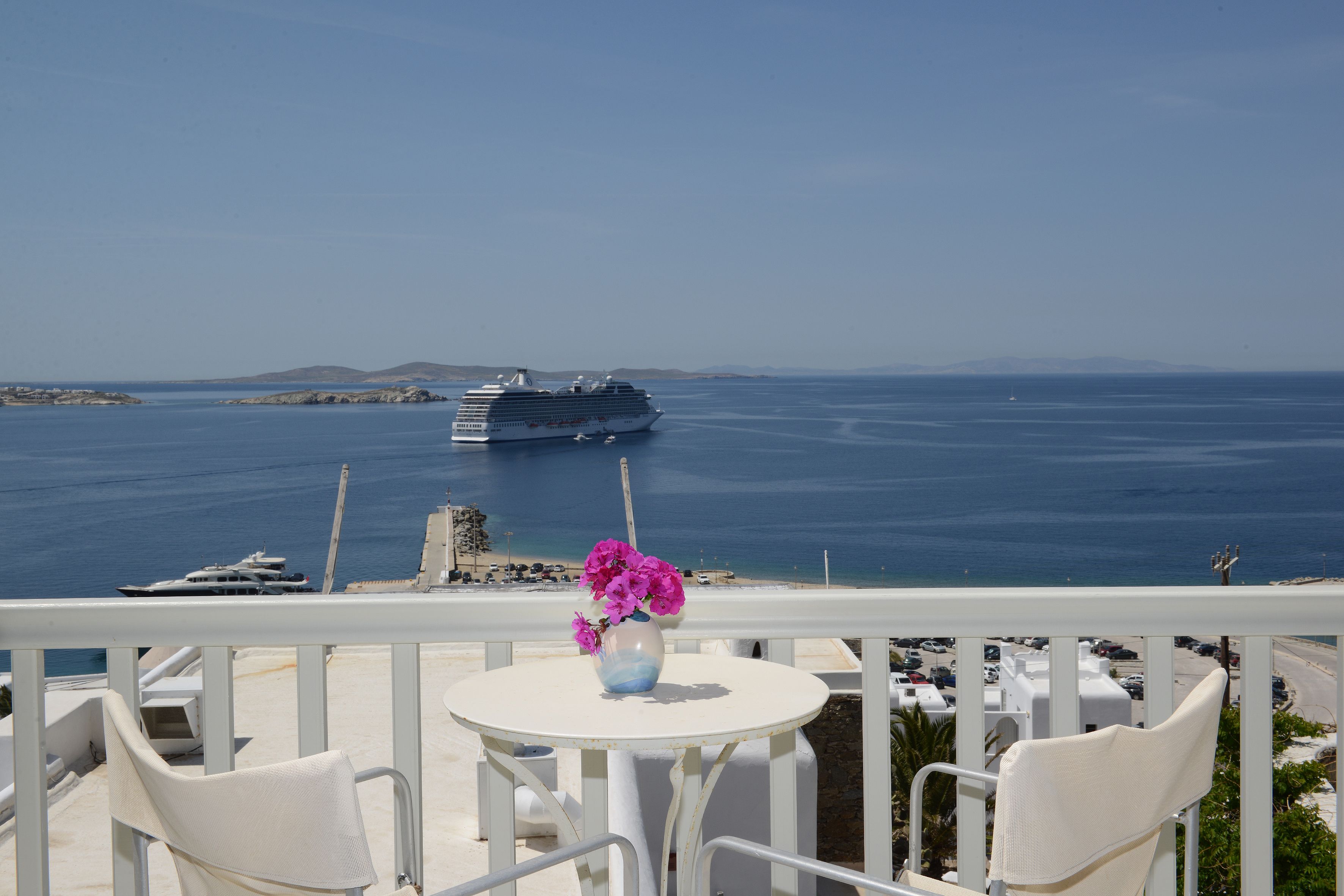 Un balcón con una mesa y sillas blancas, con vista al mar tranquilo donde se distingue un crucero en la distancia y una playa con edificios y coches.