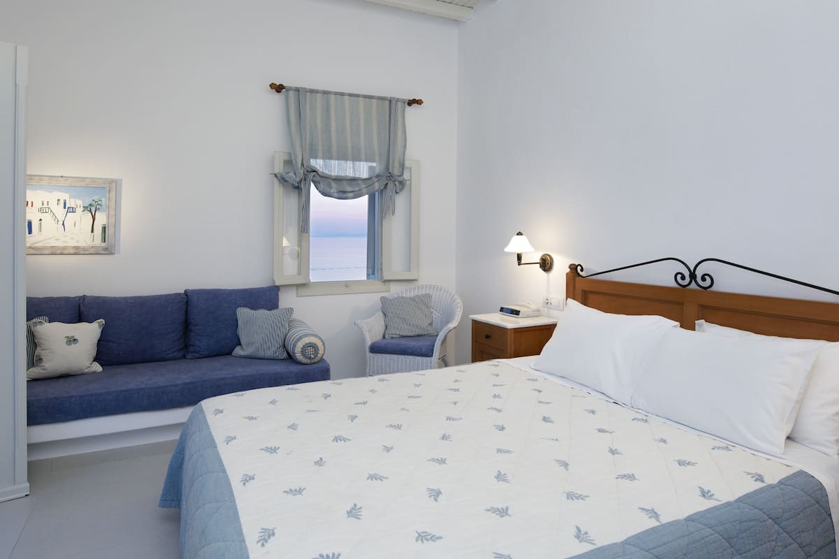 Un dormitorio con cama, sofá, sillón, mesita de noche con lámpara, ventana con cortinas y un cuadro en la pared.