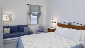 Un dormitorio con cama, sofá, sillón, mesita de noche con lámpara, ventana con cortinas y un cuadro en la pared.