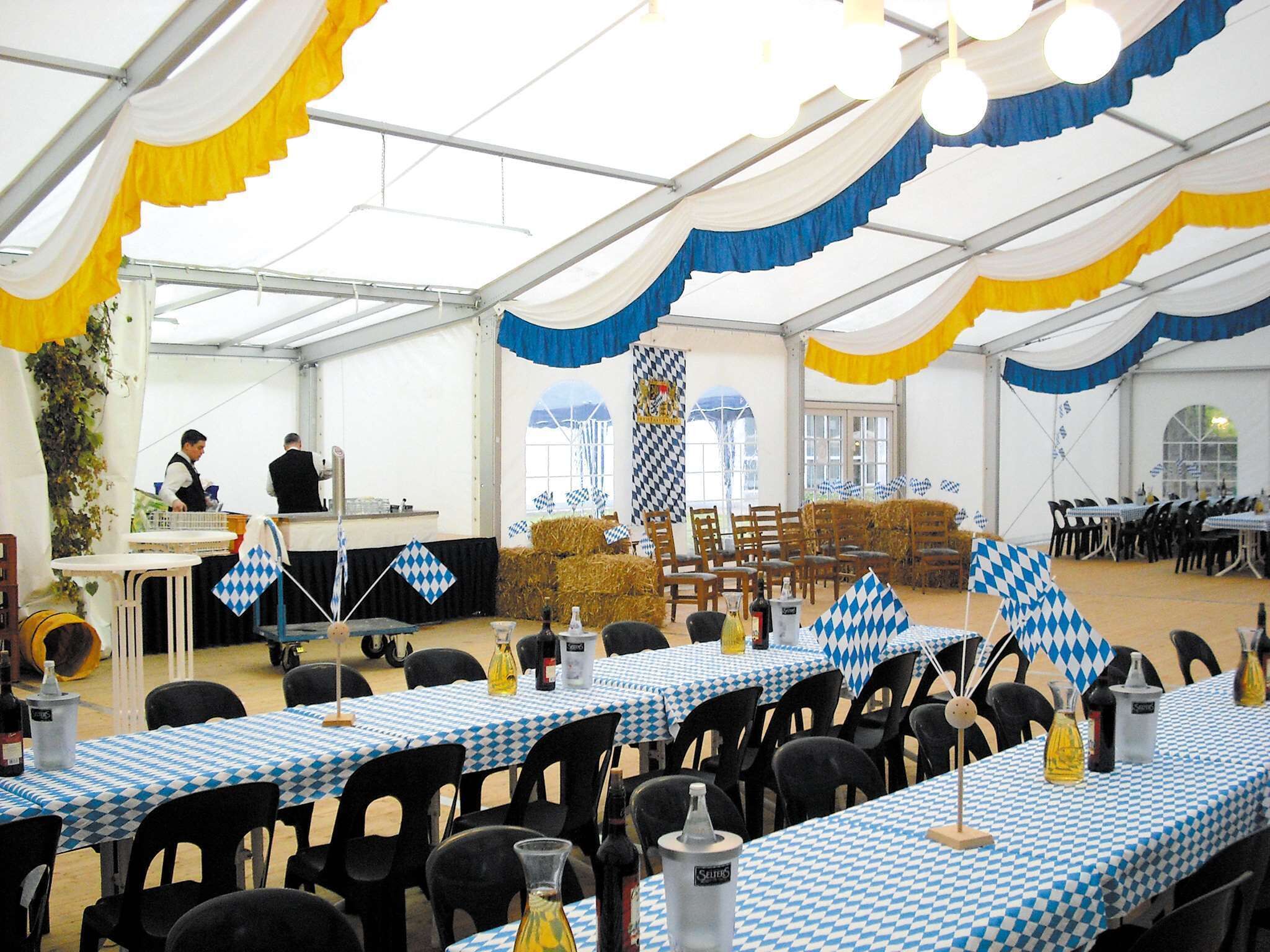 Un espacio para eventos con carpas, mesas cubiertas con manteles a cuadros, sillas y diversos objetos sobre las mesas.