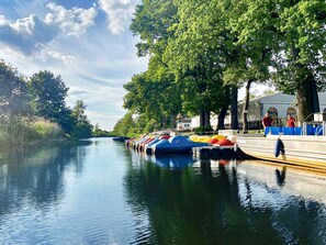 Ein ruhiger Fluss mit vertäuten Booten an der rechten Seite, ein von Bäumen gesäumter Weg und ein Gebäude mit heller Fassade.