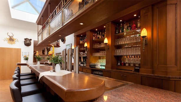 A bar area with a long counter, multiple stools, and a variety of bottles and glasses on shelves. - Morada Hotel Isetal (Gifhorn)