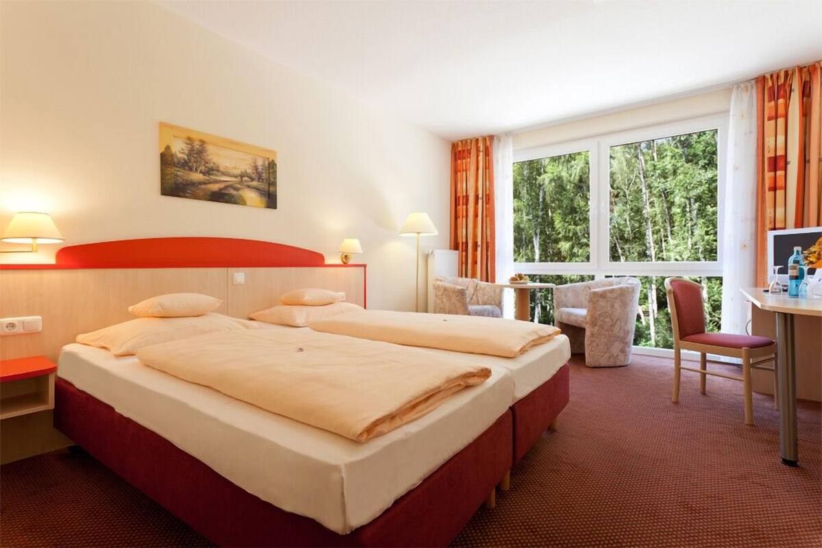 A hotel room with two beds, a desk, a chair, a lamp, and a painting on the wall.