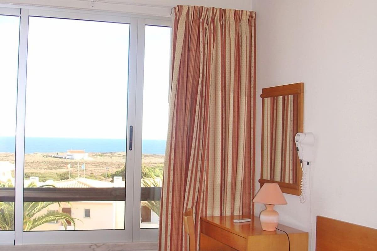 A hotel room with a bed, a desk, a chair, a lamp, a hairdryer, and a window with a view of the beach and ocean.