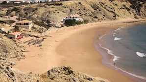 A sandy beach with a few buildings, including a large white building with a red roof and a smaller building with a red awning. - Aparthotel Navigator (Vila do Bispo)