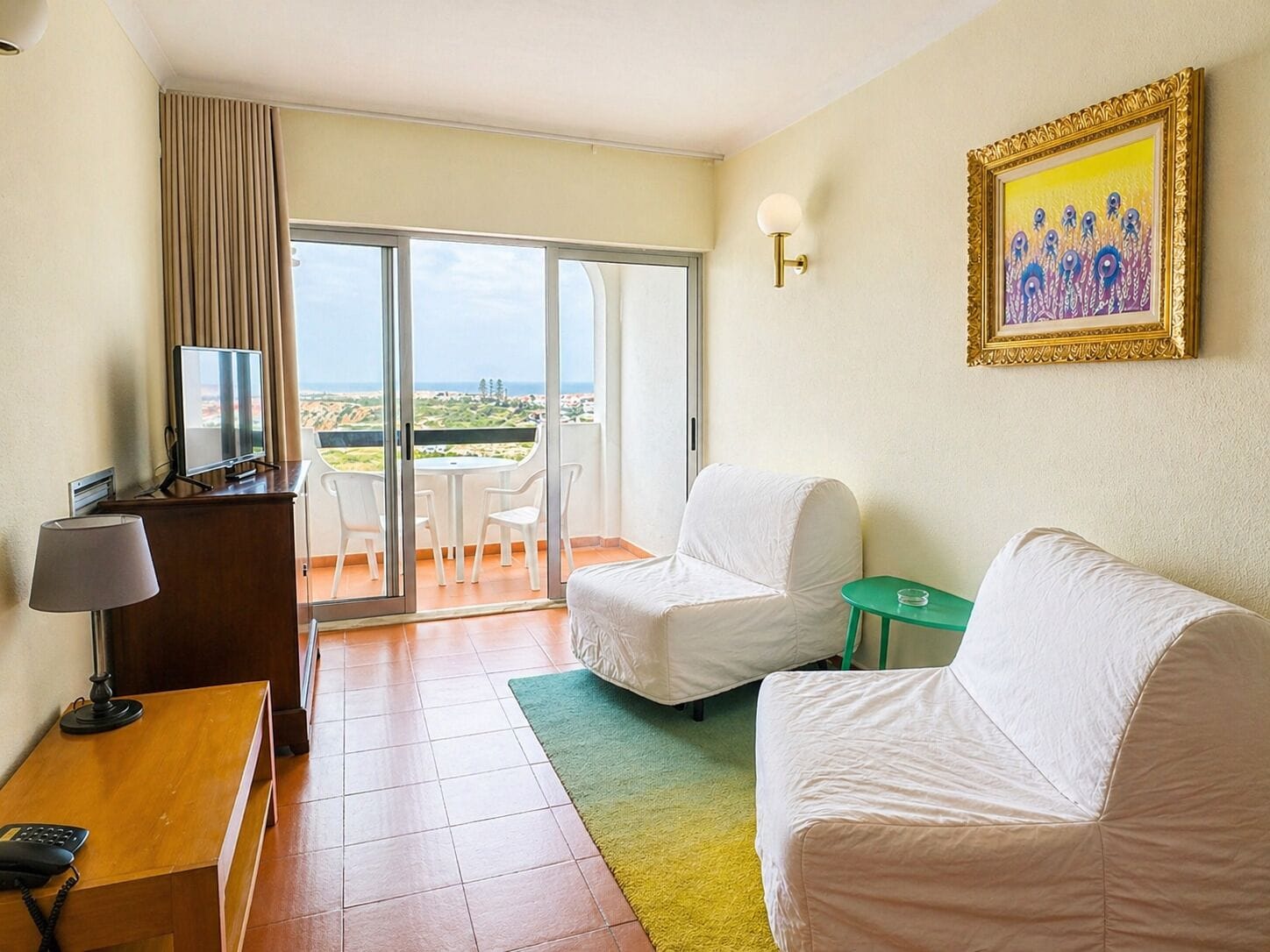 A hotel room with a balcony, a desk with a telephone, a television, and a painting on the wall.