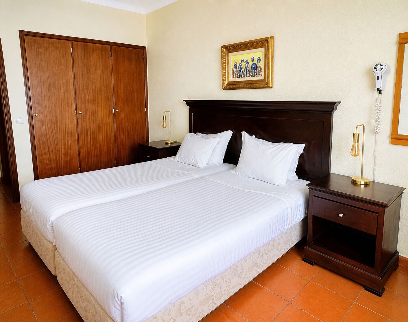 Une chambre d’hôtel avec un grand lit, une armoire en bois, des tables de chevet avec des lampes, un tableau encadré au mur et un téléphone fixé au mur.