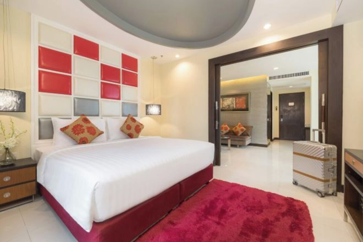 A modern hotel room with a large bed, a red accent wall, and a mirror reflecting the room.