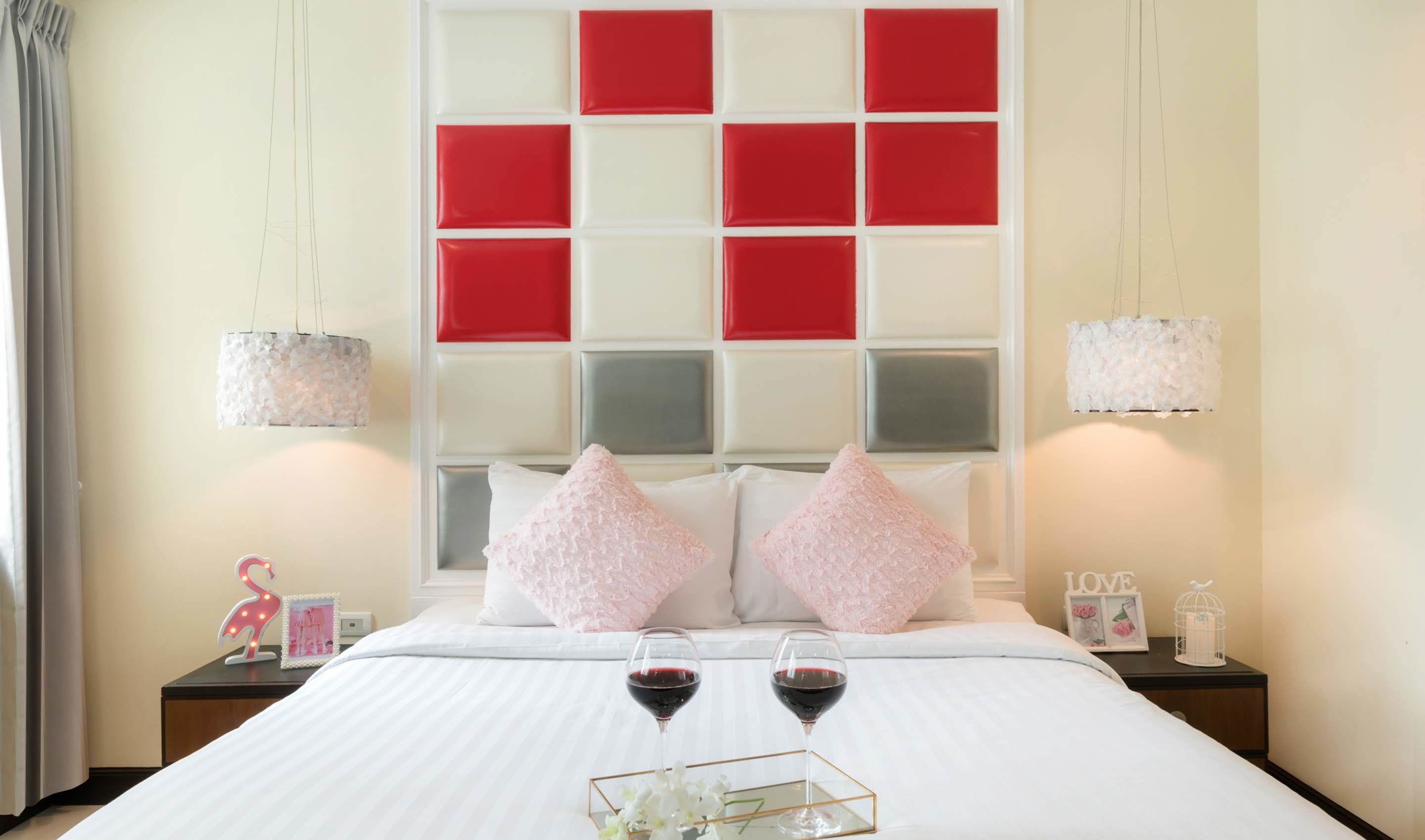 Una cama bien hecha con dos almohadas, dos mesitas de noche con lámparas y una bandeja con dos copas de vino tinto.