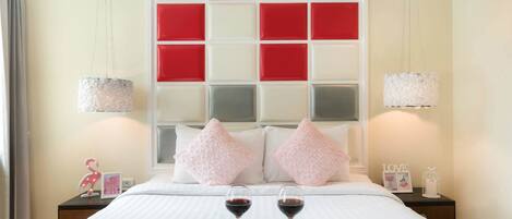 Una cama bien hecha con dos almohadas, dos mesitas de noche con lámparas y una bandeja con dos copas de vino tinto.