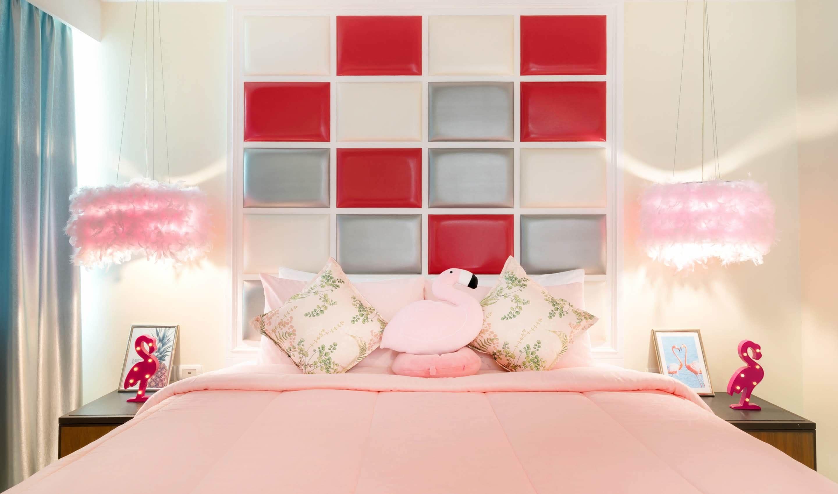 A bedroom with a pink bedspread, floral pillows, and a headboard with red and white panels.