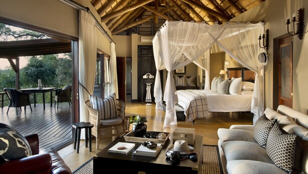 A spacious room with a large bed, a sitting area with a sofa and chairs, a dining table, and a balcony with a view of trees.