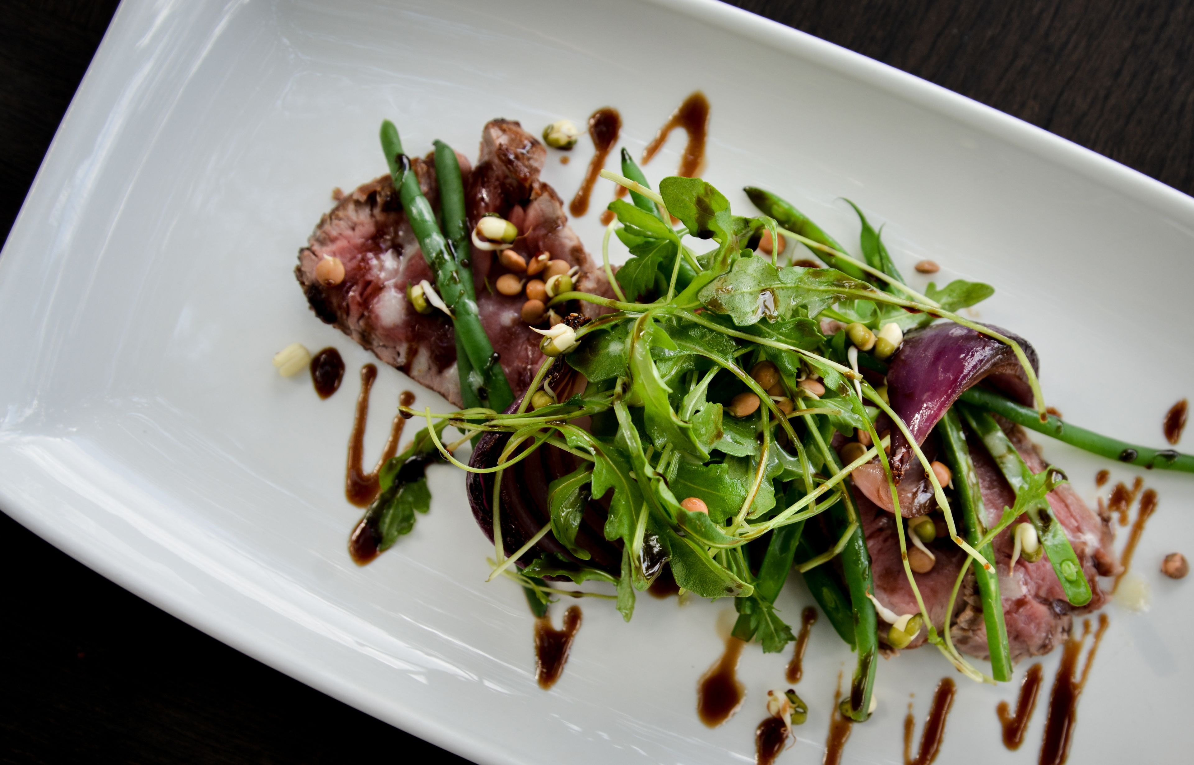 A white rectangular plate with a dish of sliced meat, green asparagus, and a salad with greens and nuts, drizzled with a dark sauce.