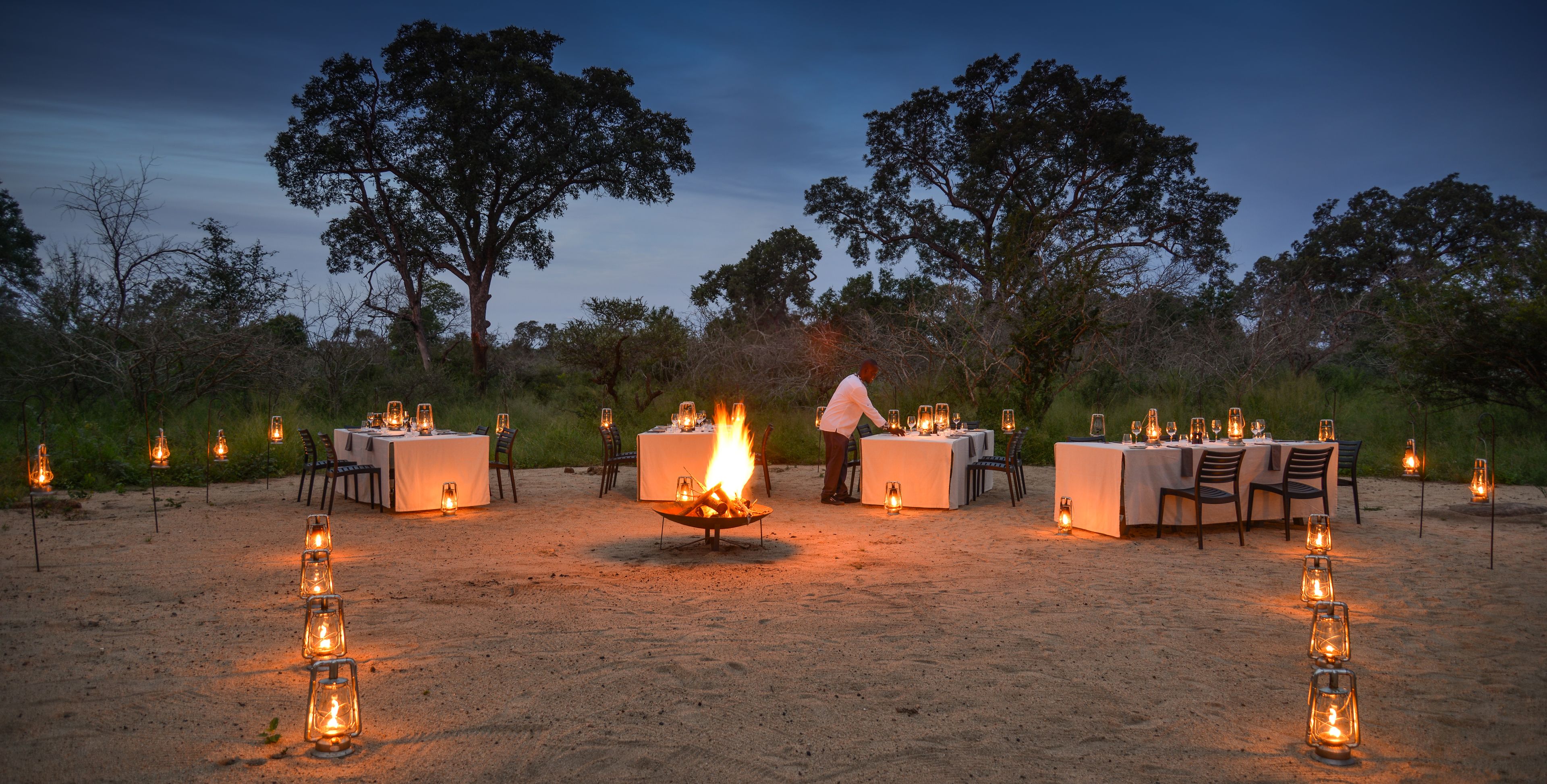 An outdoor dining setup with tables and chairs arranged around a central fire pit, surrounded by lanterns.