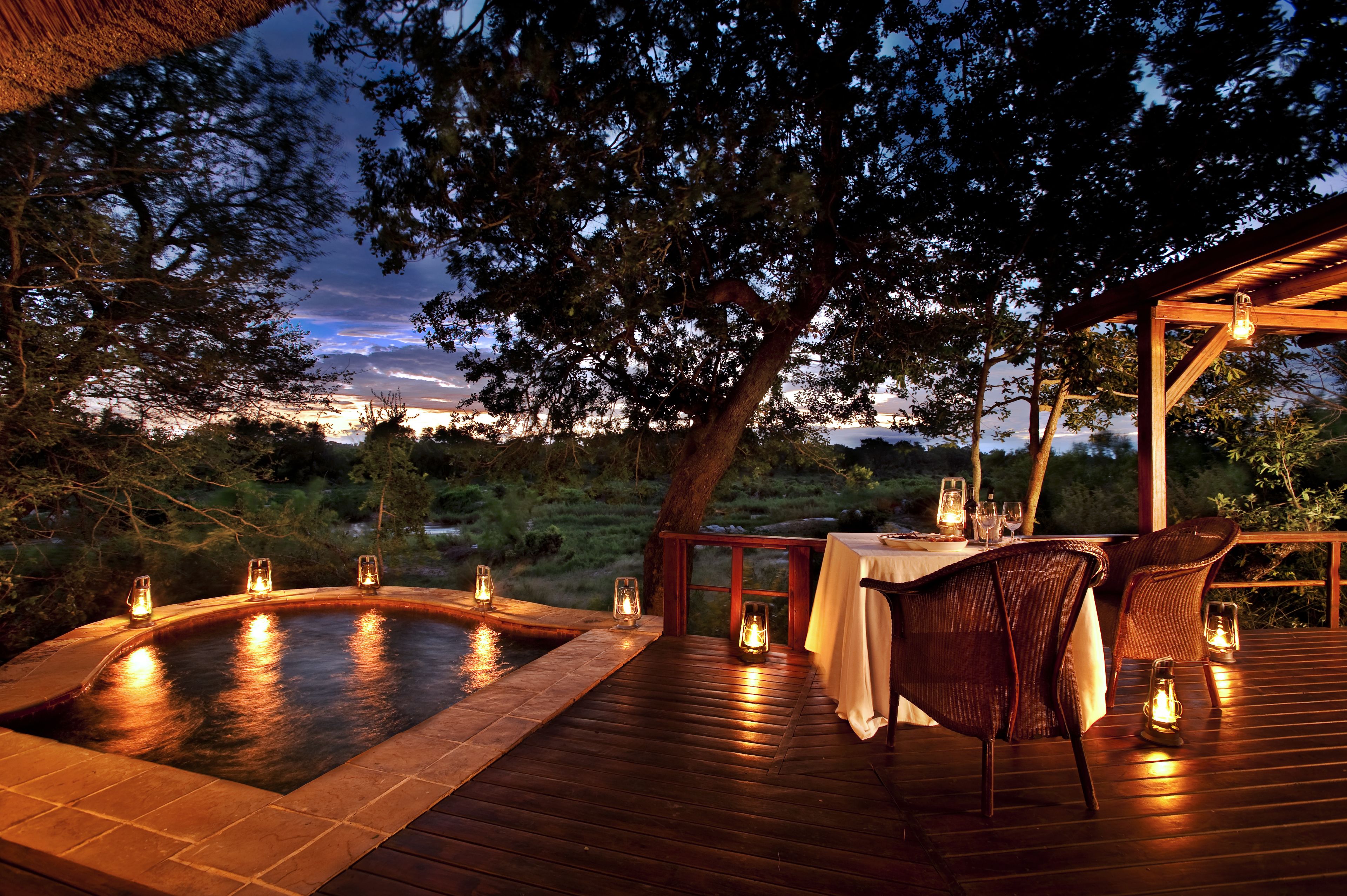 Zona de hidromasaje al aire libre con cubierta de madera, rodeada de árboles y una mesa preparada para cenar con velas.