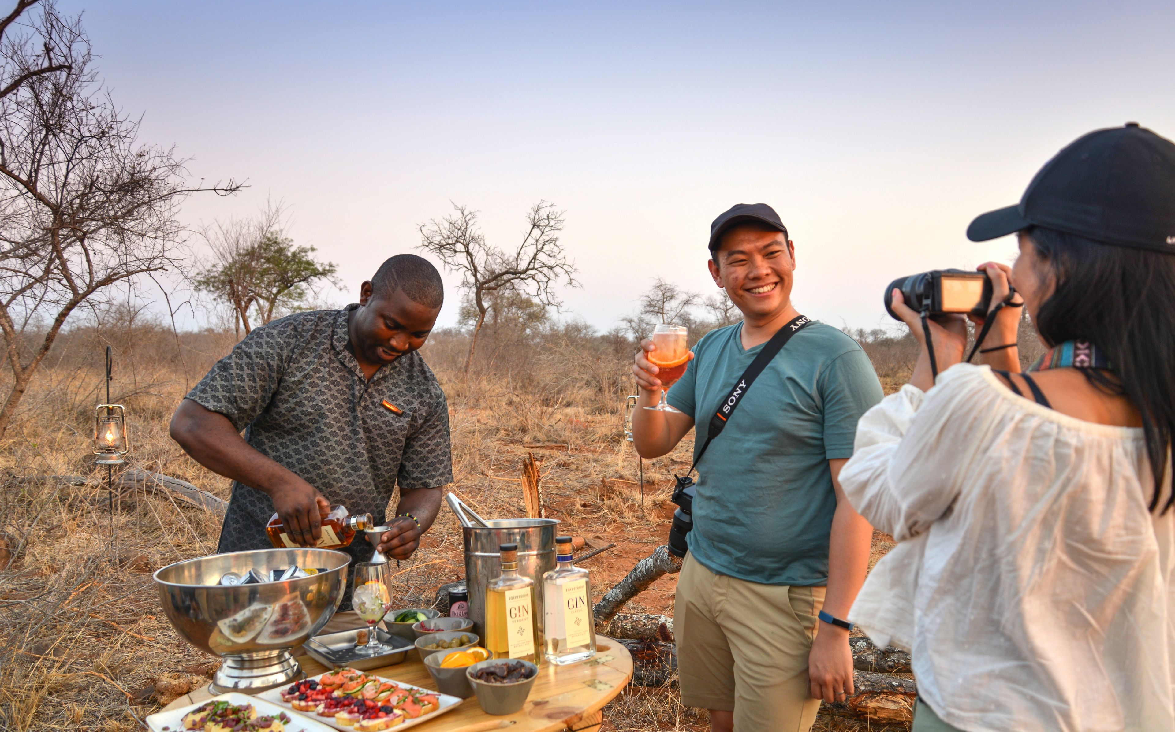 A group of people outdoors with a barbecue setup, including a metal bowl, bottles, and food.