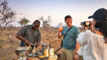 A group of people outdoors with a barbecue setup, including a metal bowl, bottles, and food.