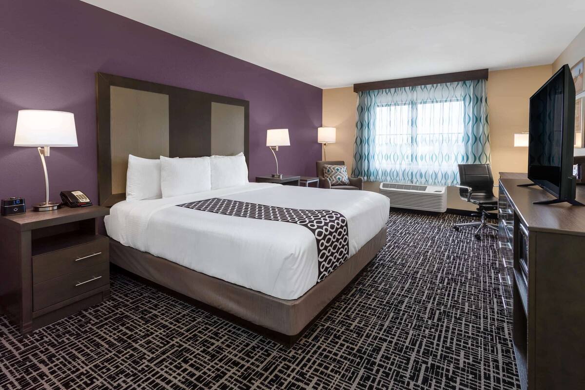 A hotel room with a large bed, two bedside tables with lamps, a desk with a chair, a television, and a window with curtains.