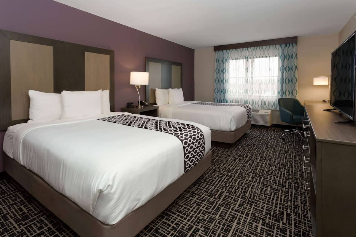 A hotel room with two beds, a desk, a chair, a TV, and a window with patterned curtains.